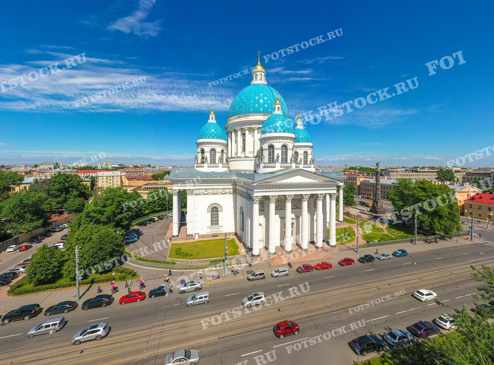 На фотографии запечатлен Троицкий собор в Санкт-Петербурге, вид с воздуха. Собор с голубыми куполами в центре кадра выделяется на фоне городской застройки под ярким солнцем и чистым голубым небом. Перед собором проходит оживлённая дорога, машины движутся по дорогам, вдоль которых расположены зелёные деревья. Эта фотография может быть использована в туристических буклетах и гидах, в качестве иллюстративного материала в статьях об архитектуре и истории города, а также может служить украшением в интерьере или для создания почтовых открыток.